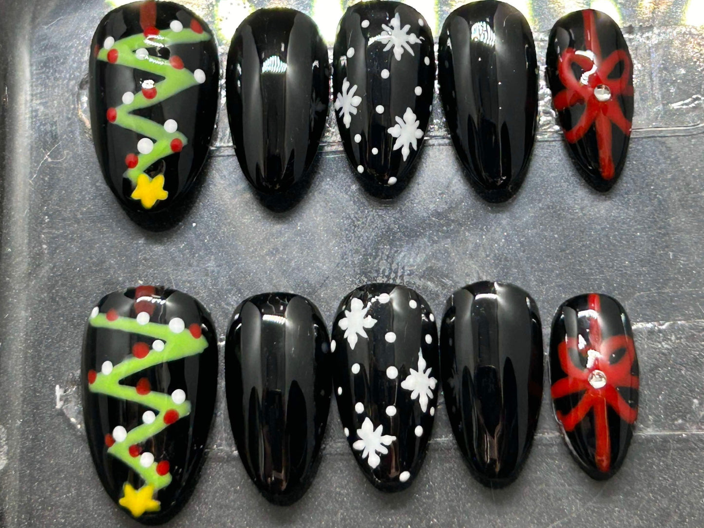 A festive set of glossy gel press-on nails featuring Christmas trees, snowflakes, and red gift bows with rhinestone accents. Displayed in a clear case, the nails come in various sizes and shapes, perfect for holiday glam.