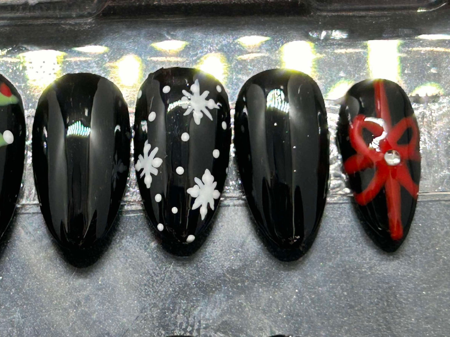 A festive set of glossy gel press-on nails featuring Christmas trees, snowflakes, and red gift bows with rhinestone accents. Displayed in a clear case, the nails come in various sizes and shapes, perfect for holiday glam.
