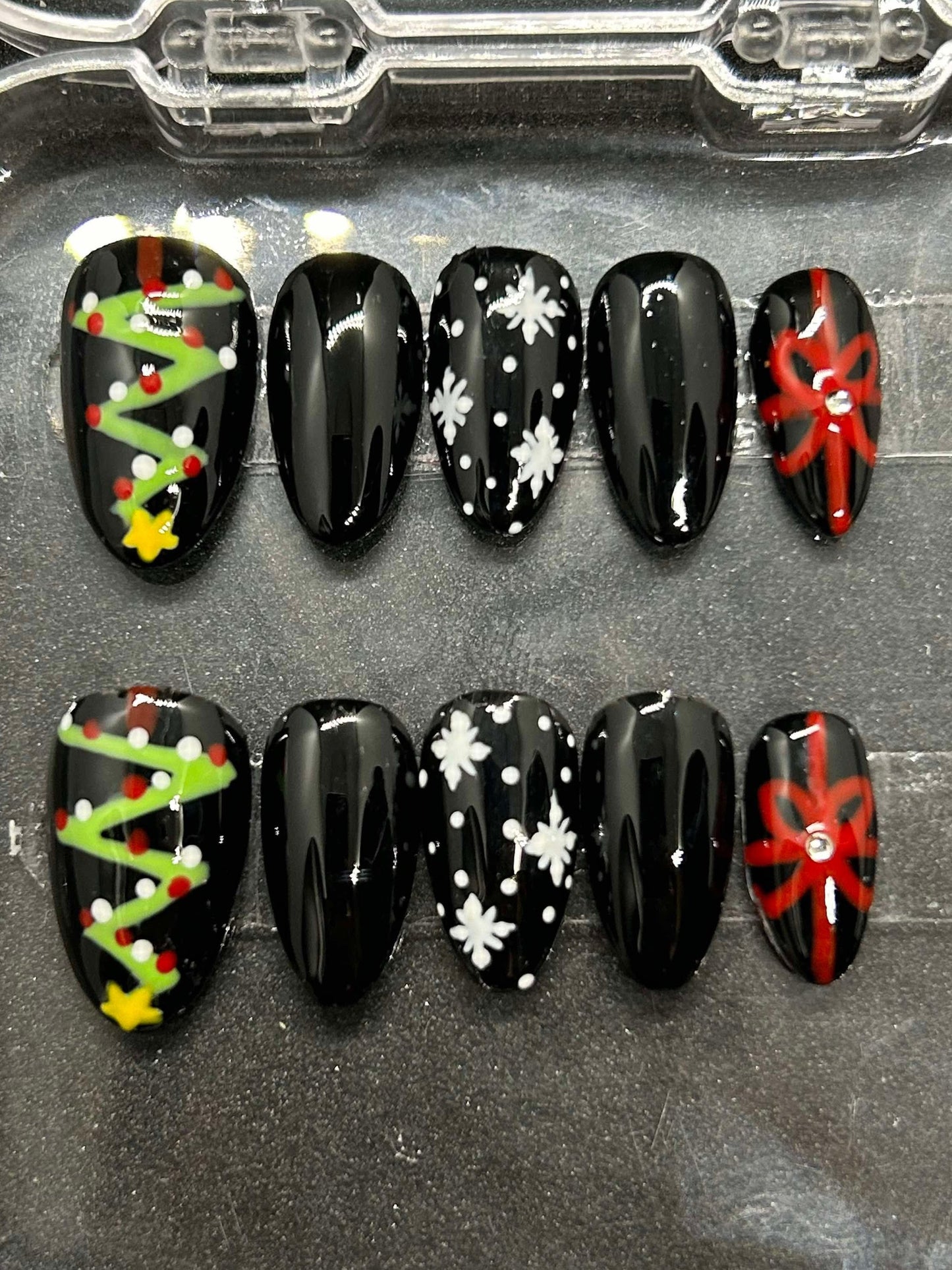 A festive set of glossy gel press-on nails featuring Christmas trees, snowflakes, and red gift bows with rhinestone accents. Displayed in a clear case, the nails come in various sizes and shapes, perfect for holiday glam.