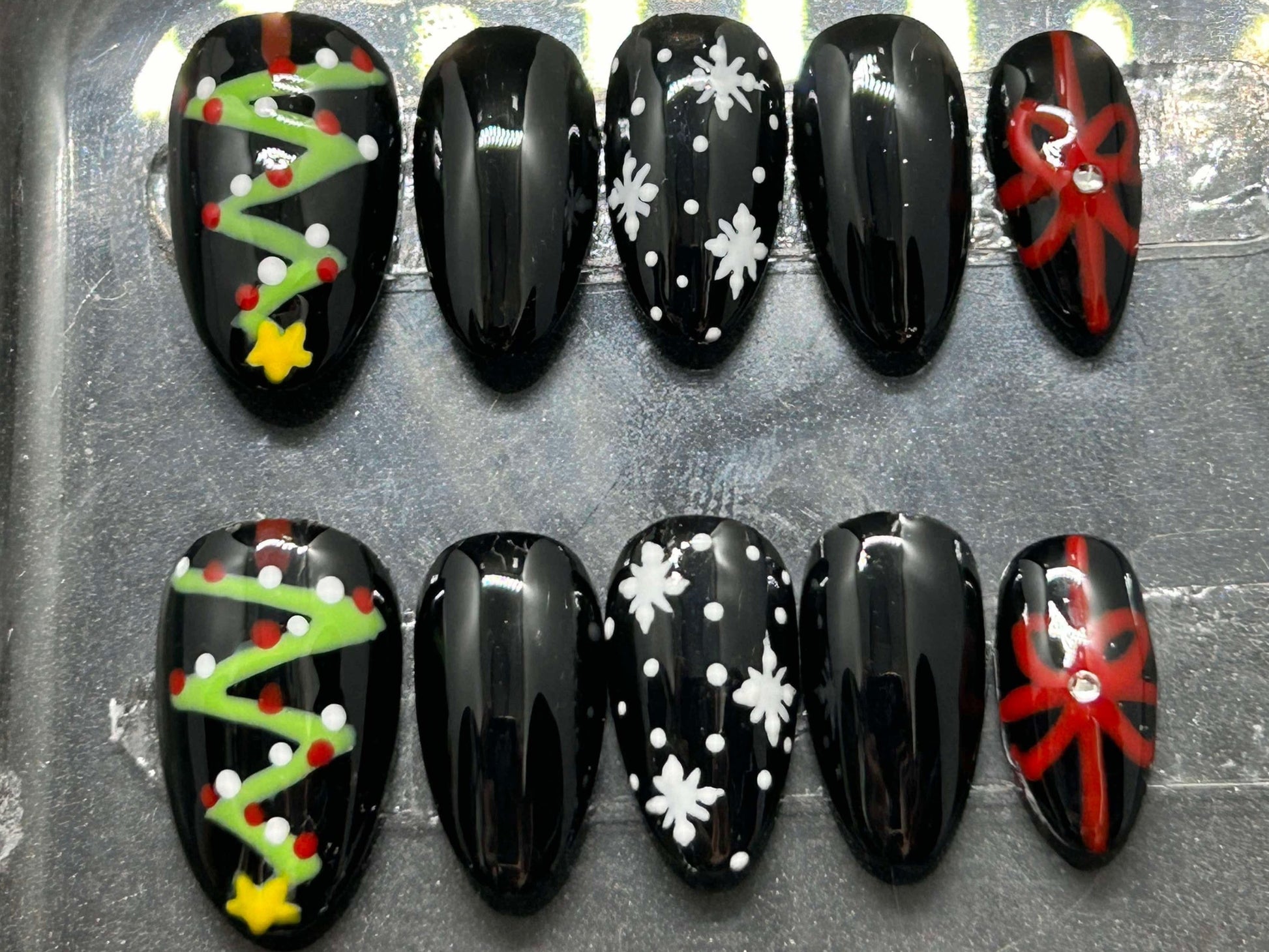 A festive set of glossy gel press-on nails featuring Christmas trees, snowflakes, and red gift bows with rhinestone accents. Displayed in a clear case, the nails come in various sizes and shapes, perfect for holiday glam.